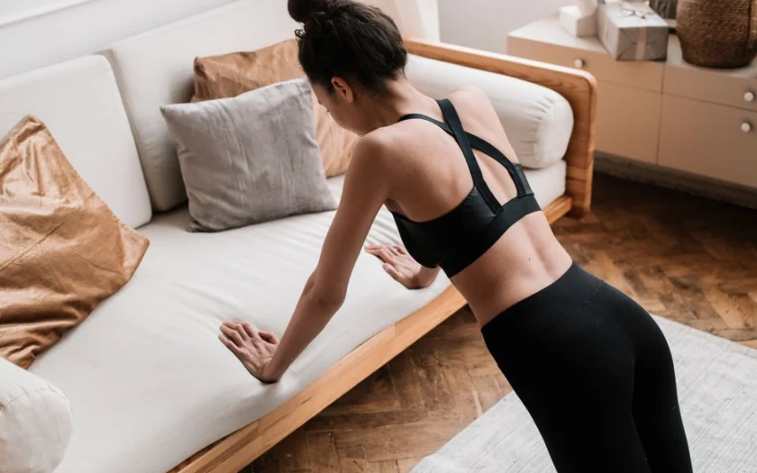 An active adult woman performs push-ups indoors using a sofa for support, emphasizing a healthy lifestyle.