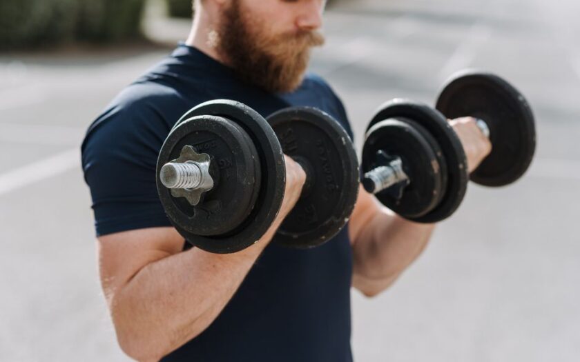 Crop sporty male making effort doing exercise with dumbbells while working out on blurred background of sports ground